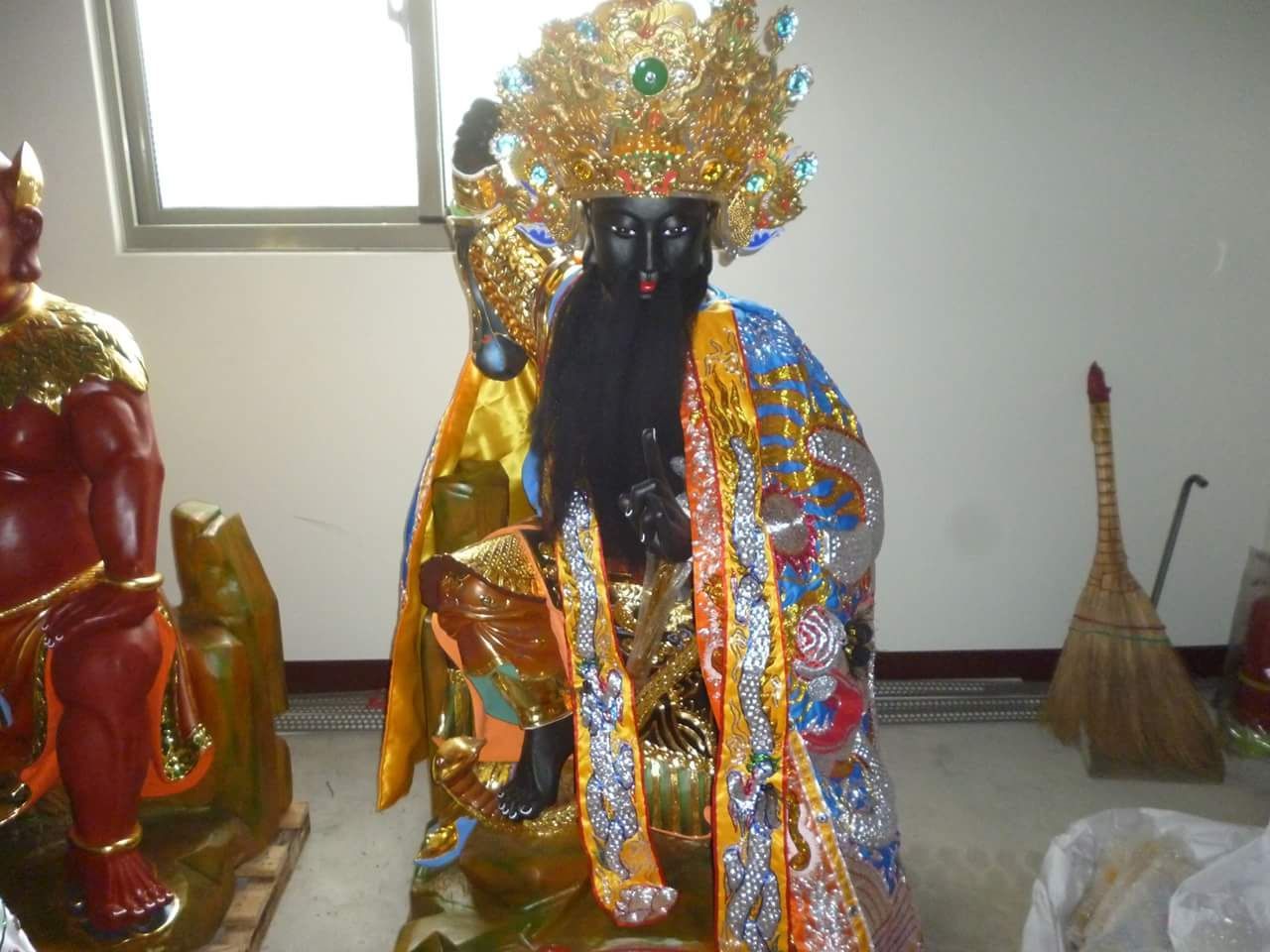 一尊裝飾華麗的黑色雕像,身穿金藍色長袍,頭戴頭飾,擺放在室內窗邊,旁邊放著一把掃帚。