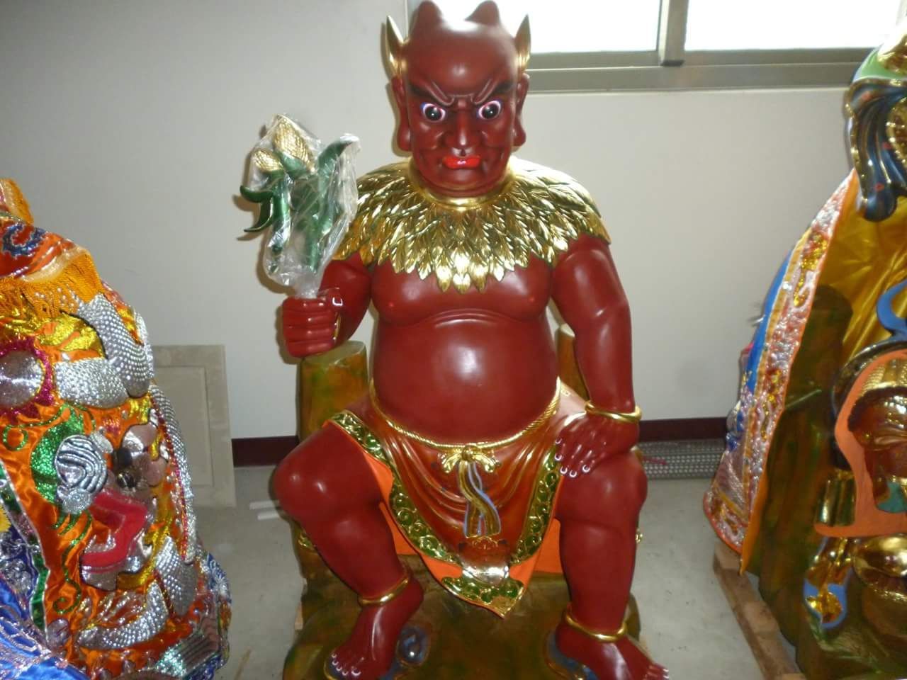 一尊紅角雕像,佩戴金項圈和飾物,端坐於室內,周圍環繞著色彩繽紛的裝飾人物。