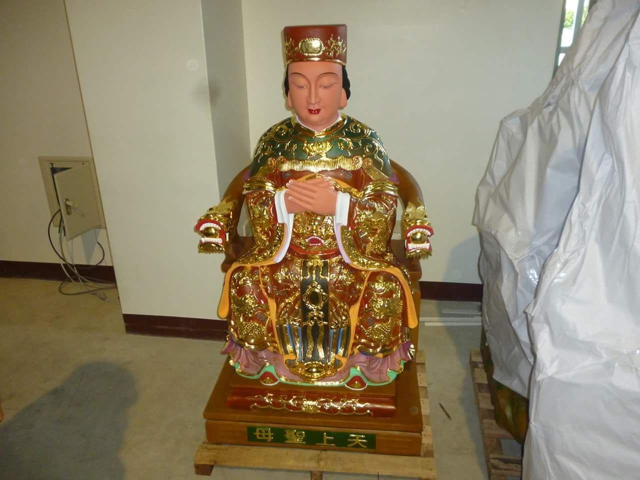 室內房間裡,一尊色彩鮮豔的坐像立於木質底座上,飾以金色和紅色細節。
