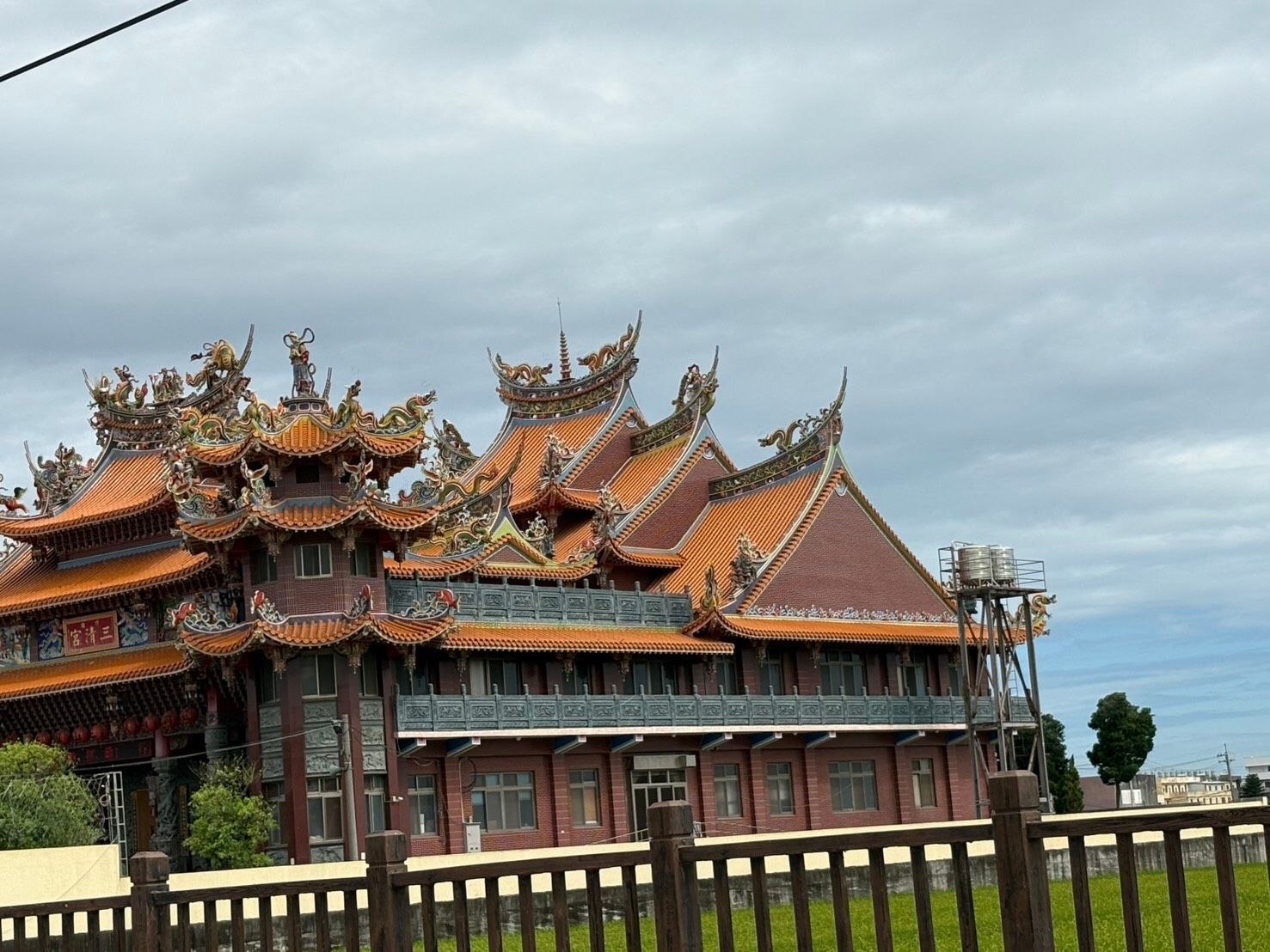 在陰雲密佈的天空下,一座有著橘色屋頂的華麗寺廟矗立在道路和開闊的田野旁。