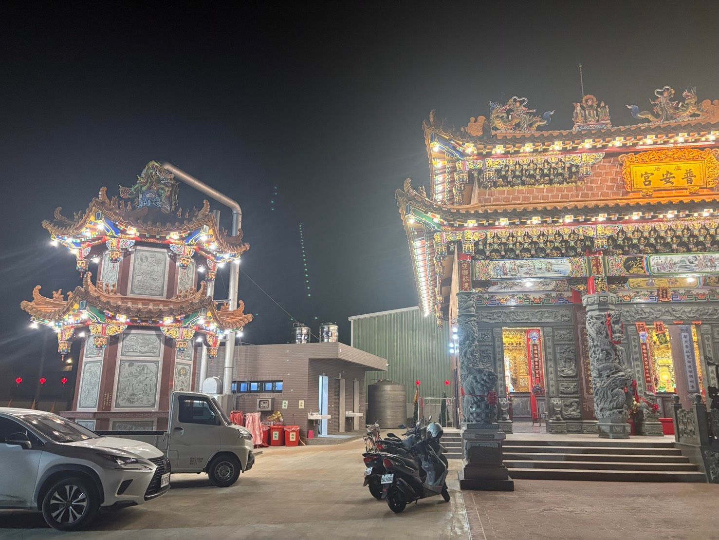 夜幕降臨,寺廟庭院燈火輝煌,裝飾華麗的建築群、停放的車輛和一輛摩托車映入眼簾。