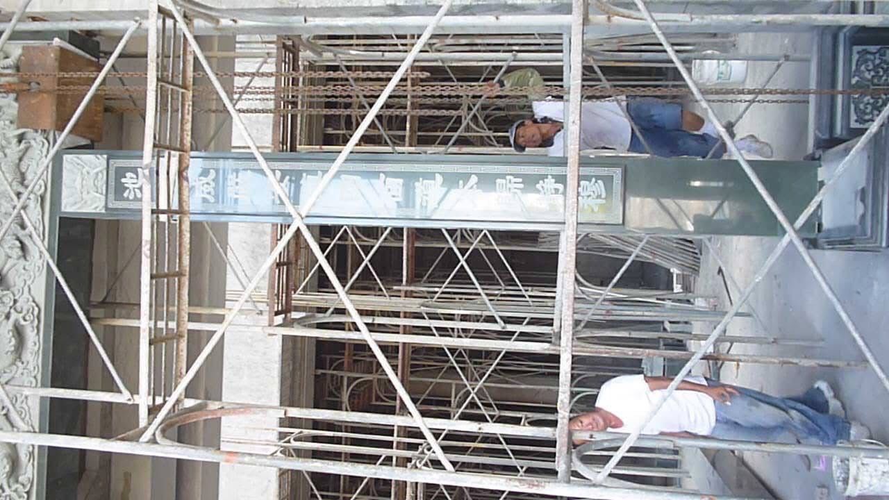 建築工人在建築物外牆旁的鷹架上工作,鷹架有金屬框架和藍色遮蓋物。