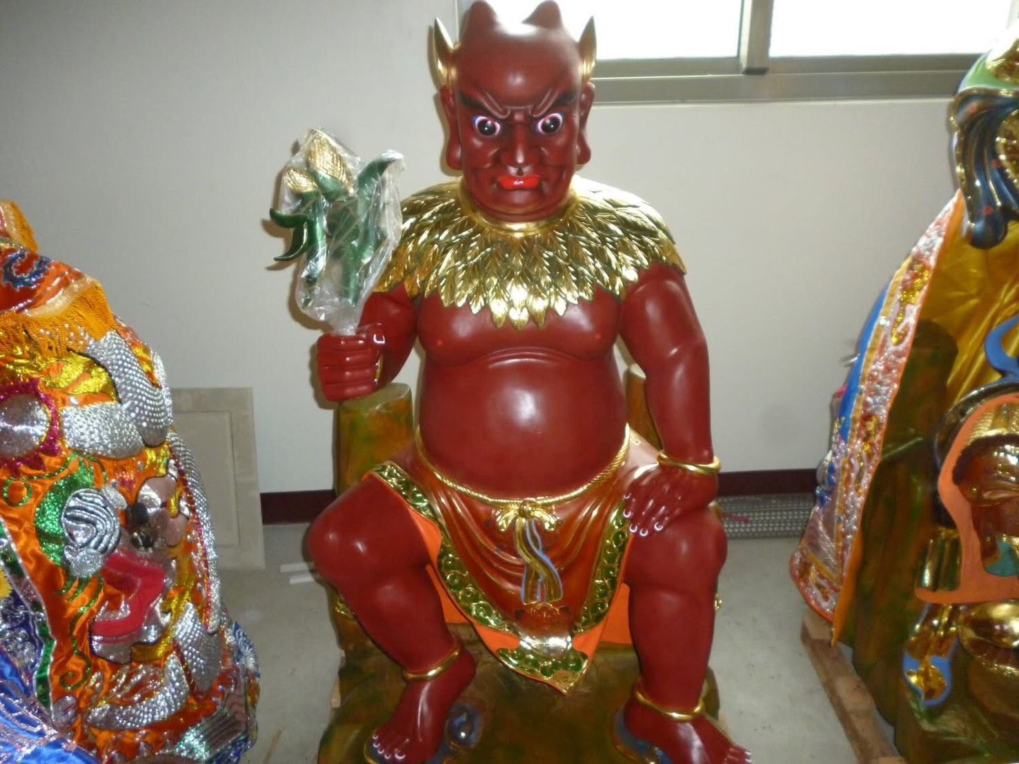 紅色惡魔雕像坐在凳子上,手裡拿著一個小物件,兩側各有一個裝飾華麗的人物雕像。