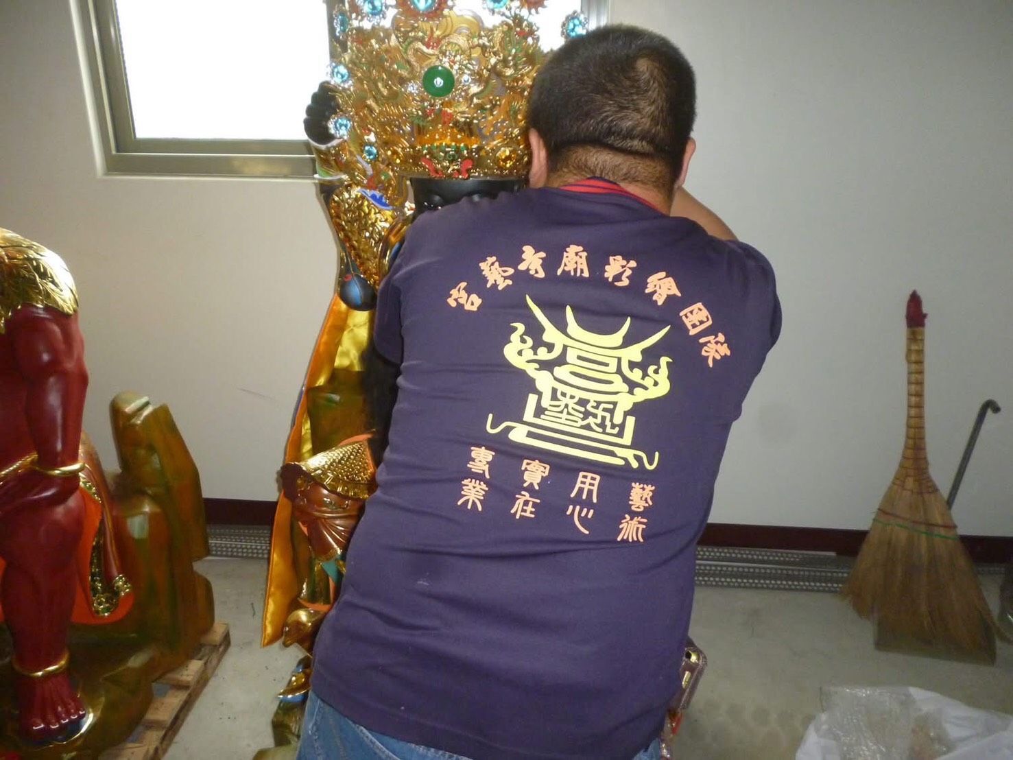 身穿紫色襯衫的人跪在神龕內的一尊金色雕像前,旁邊放著一把掃帚。