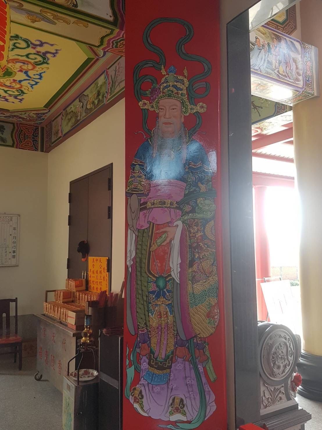 室內祭壇旁,一根紅色柱子上繪有色彩繽紛的寺廟壁畫,陽光灑在門口。