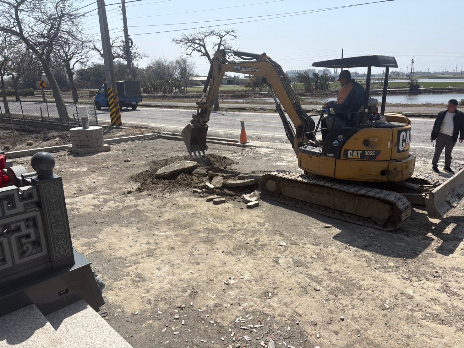 一輛黃色挖土機正在路邊建築工地挖溝,附近有工人在作業。