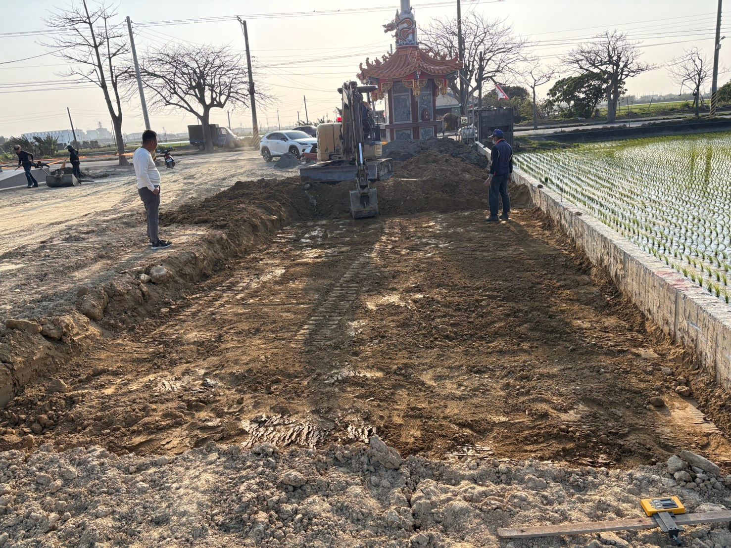 工人們在一片被水淹沒的稻田旁泥濘的道路上作業,前方中央有一座小神社。
