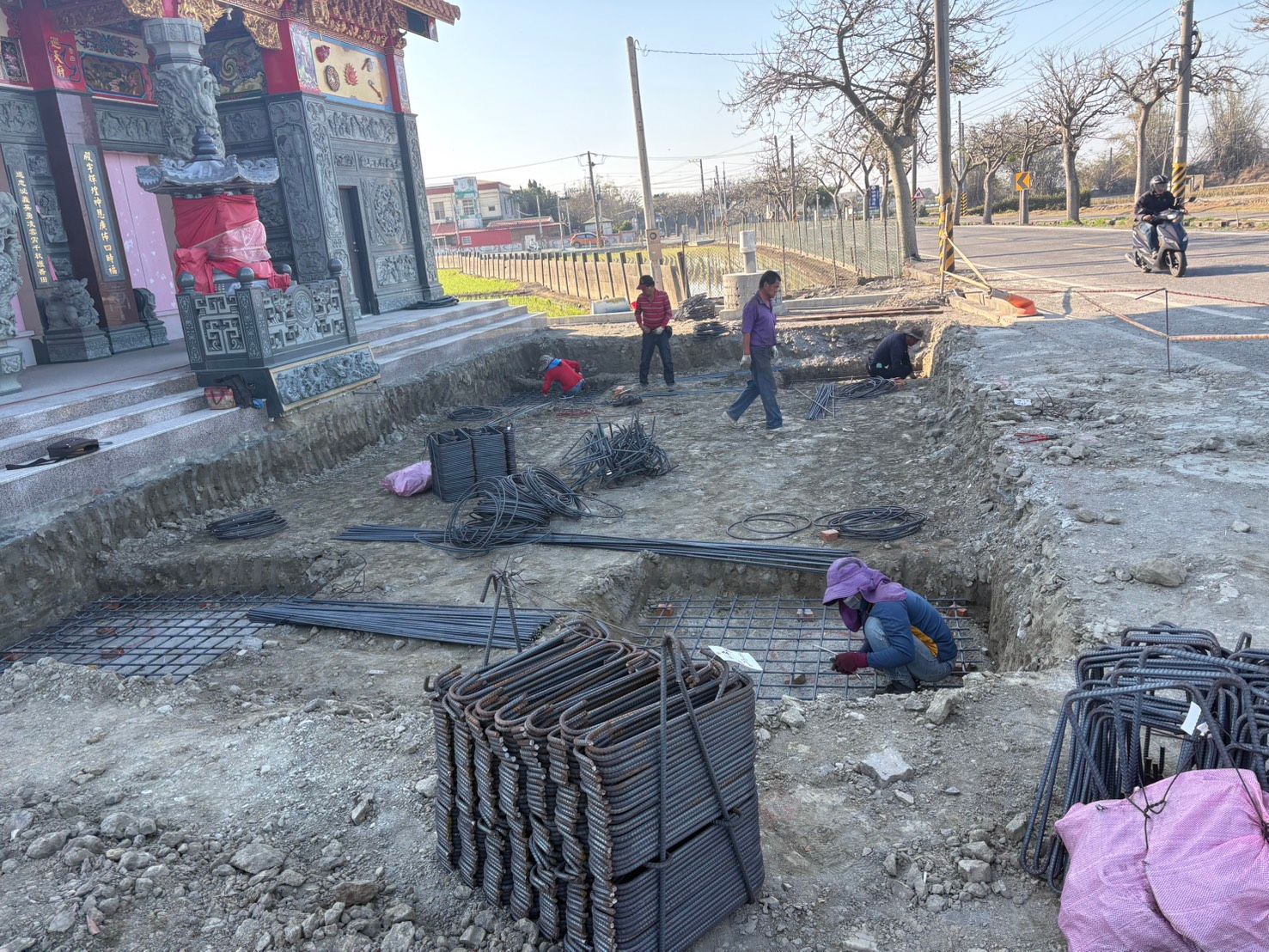 建築工人在磚砌建築和停放的摩托車旁的土路邊鋪設鋼筋。