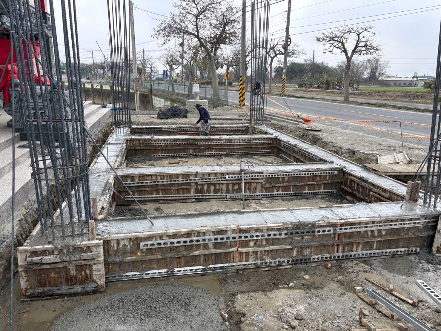 路邊建築工地,鋼筋、混凝土模板,一名工人在潮濕陰沉的路面附近作業。