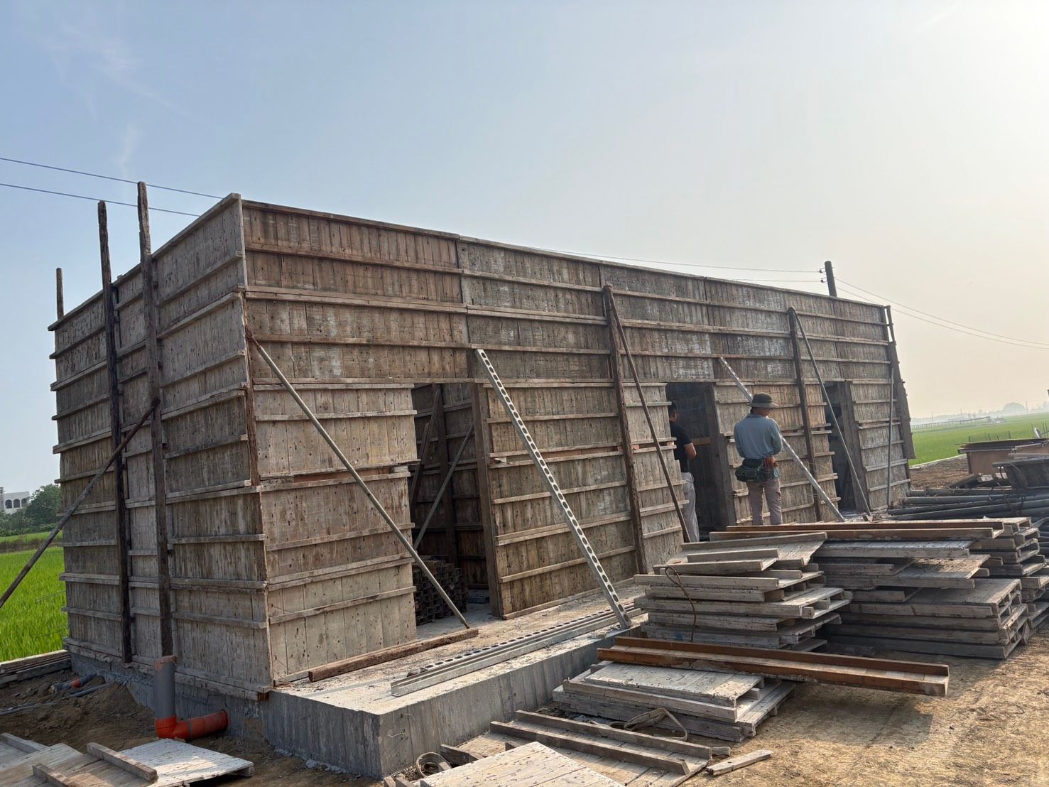 鄉村建築工地上,一座半成品混凝土結構,有木製模板,兩名工人正在施工。