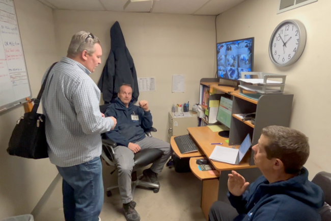 Two men are talking in a room with a clock on the wall