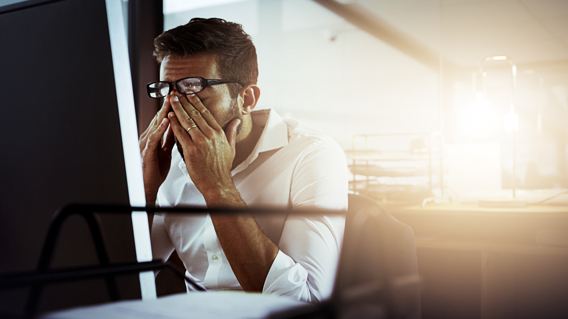 A man is covering his face with his hands while looking at a computer screen.