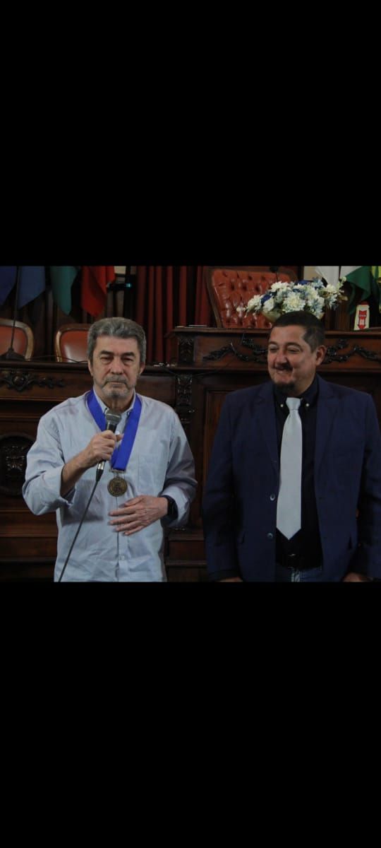 Dois homens estão de pé, um ao lado do outro, em uma sala, e um deles usa uma medalha pendurada no pescoço.