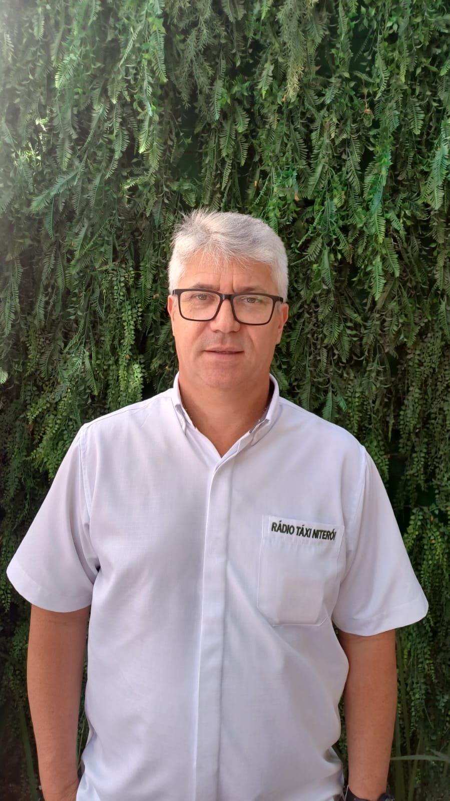 Um homem usando óculos e uma camisa branca está parado em frente a uma árvore.