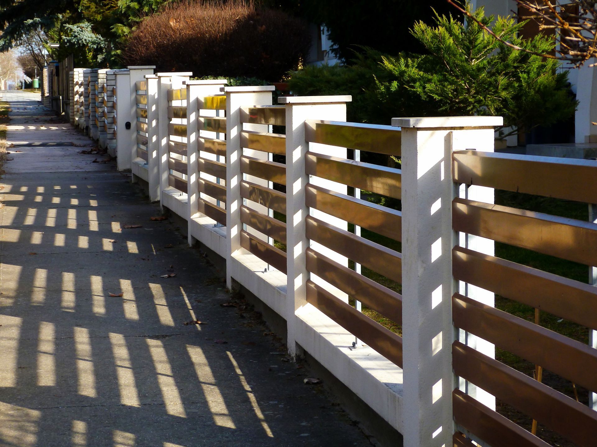 Fence with horizontal wooden slats between white concrete posts casting shadows on the sidewalk.