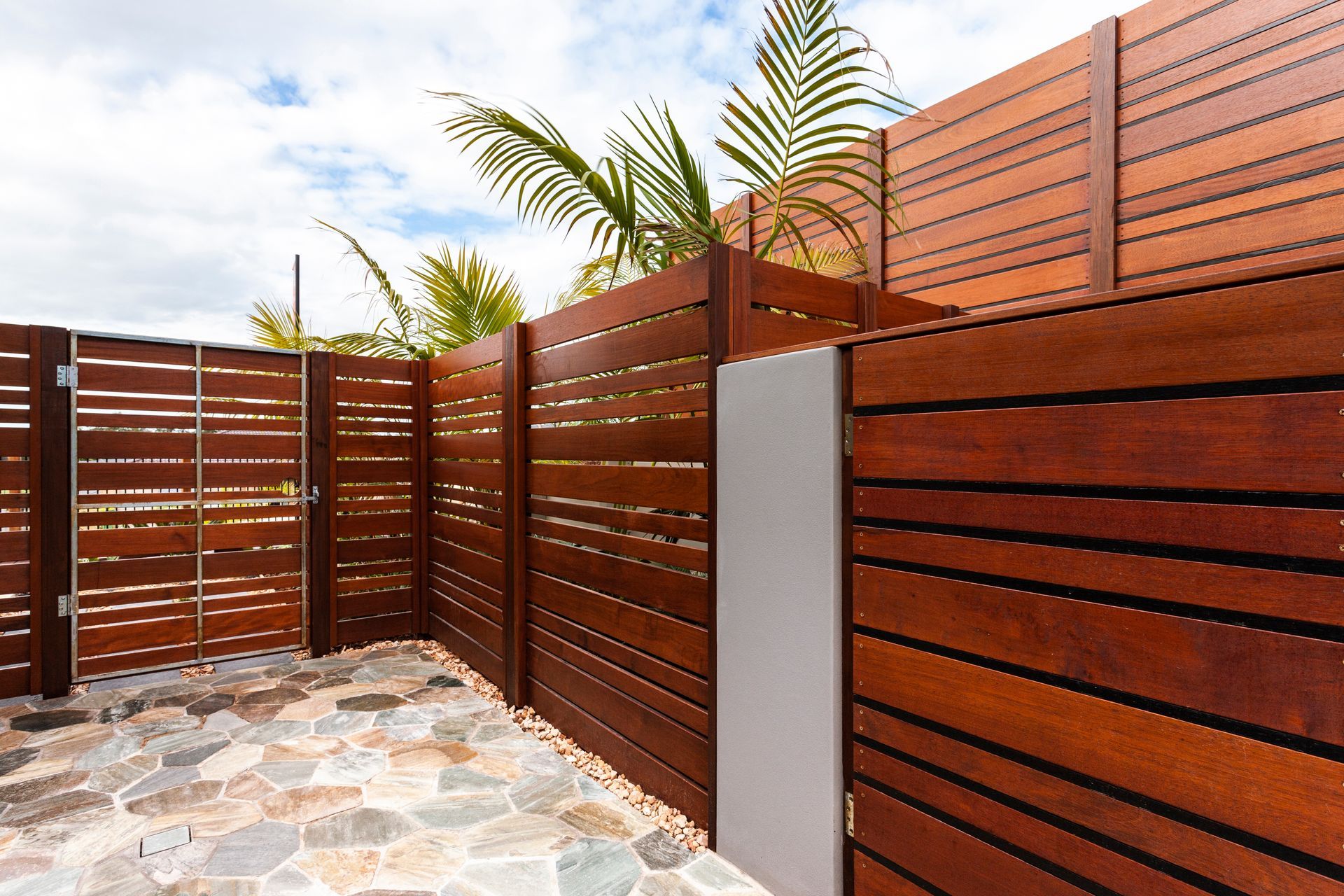 Wooden fence with horizontal slats, gate, stone ground, and palm fronds.