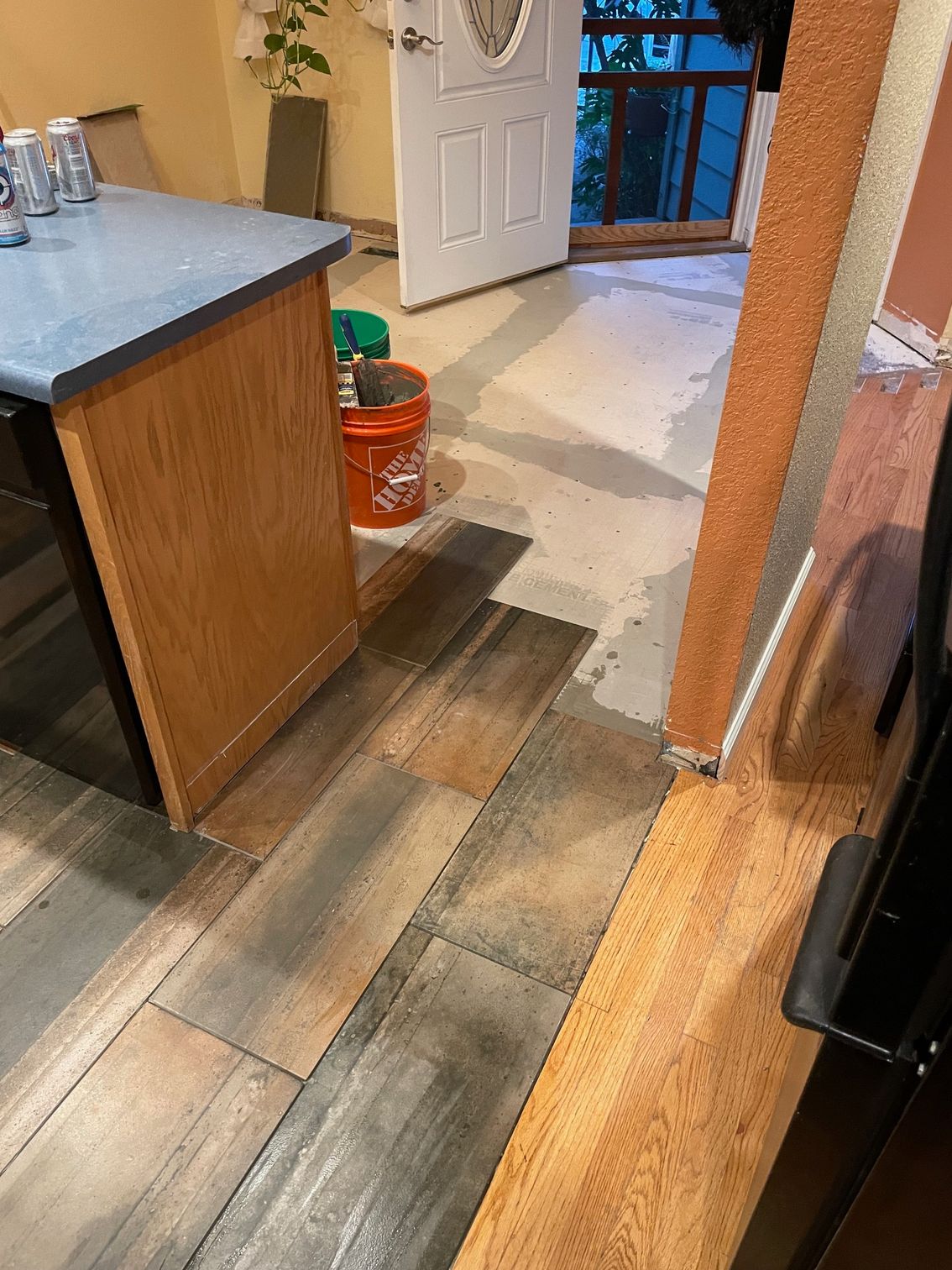 Wooden floor with varying colors meets concrete. A kitchen cabinet and open door are visible.
