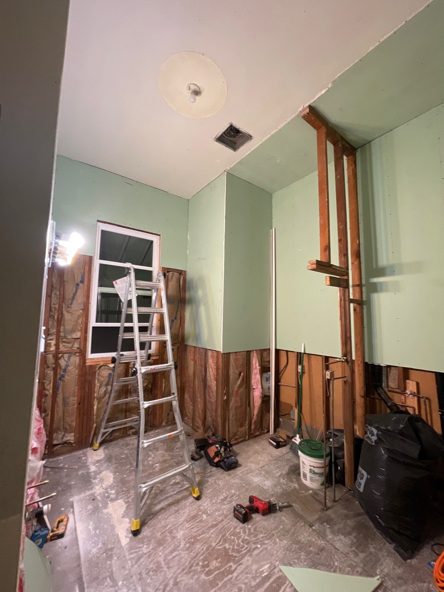 Room undergoing renovation, exposed studs, insulation visible. Ladder, tools, and partial walls. Pale green walls, white window.
