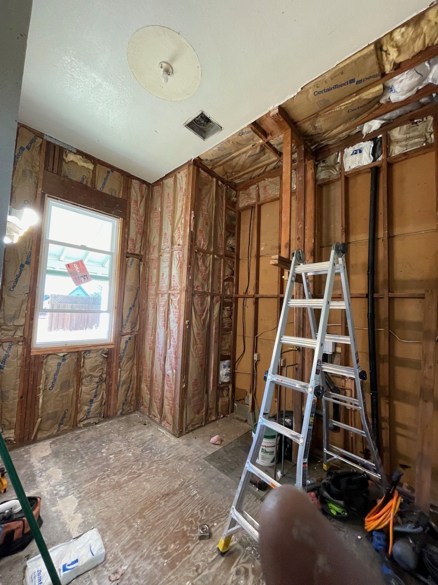 Interior room gutted, insulation exposed, ladder in front, window and ceiling light visible.