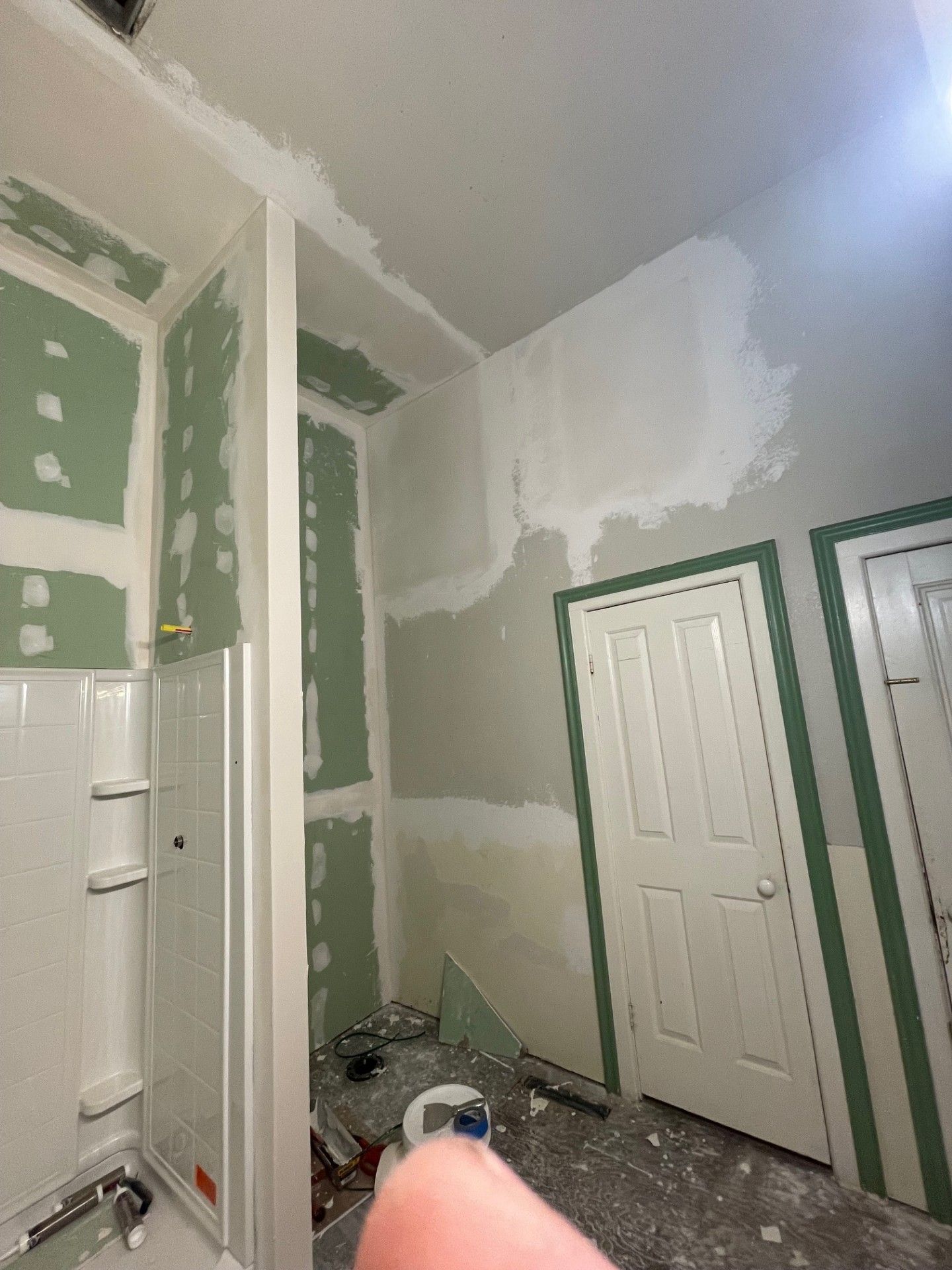 Bathroom under construction; green drywall, white trim, partially plastered walls, shower stall, white doors, tools on floor.