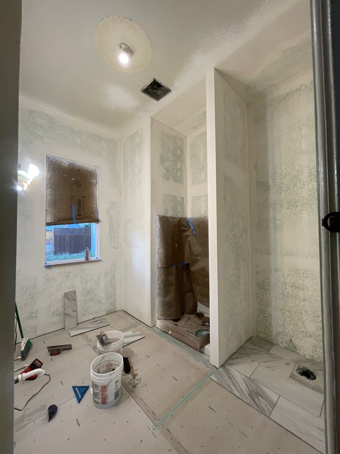 Bathroom under renovation, drywall installed, unfinished surfaces, tools on floor, window.