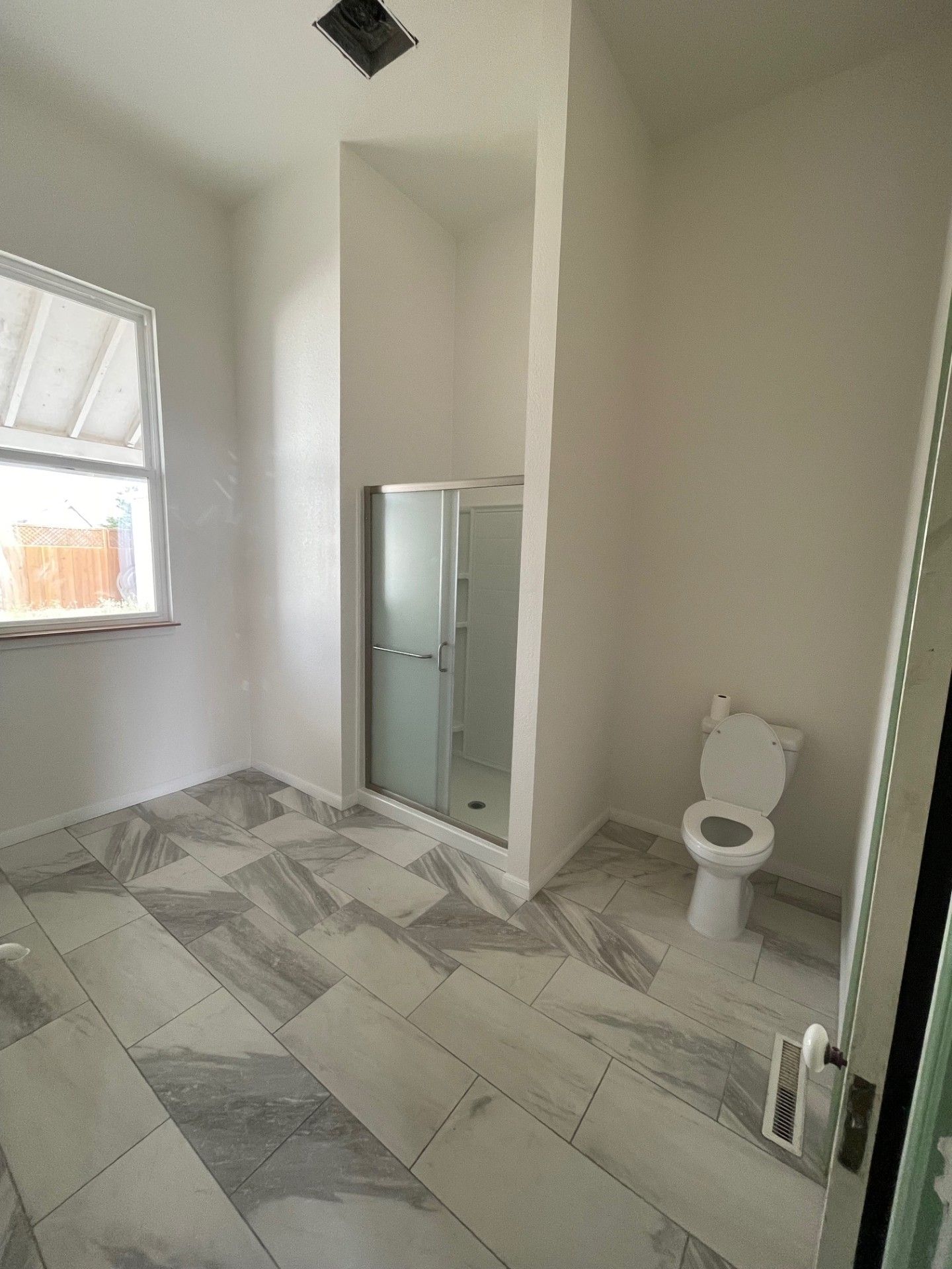 Bathroom with a tiled floor, shower stall, toilet, and window. Walls are white.