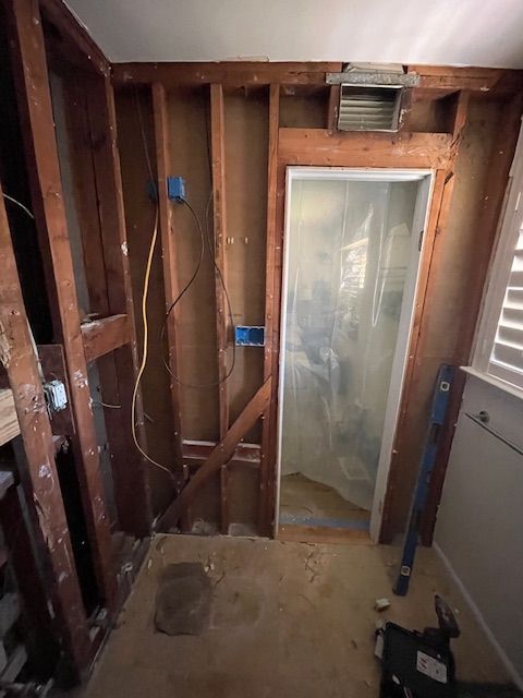 Bathroom undergoing renovation: exposed wood framing, electrical wiring, plastic-covered shower entrance, and scattered debris on the floor.