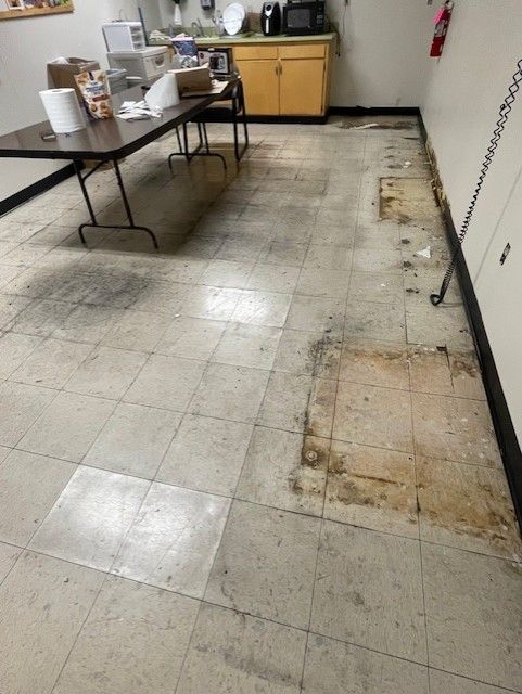 A worn, dirty tile floor in a room with a table, cabinets, and appliances. Some tiles are missing.