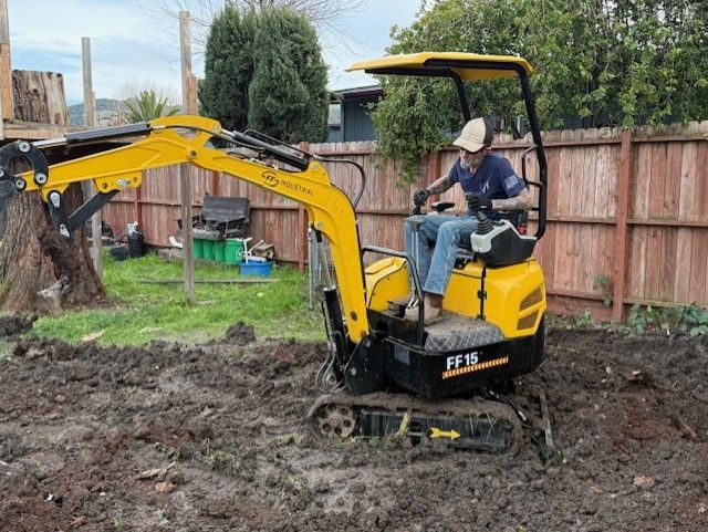 Person operating a yellow mini-excavator in a muddy yard near a wooden fence and trees.