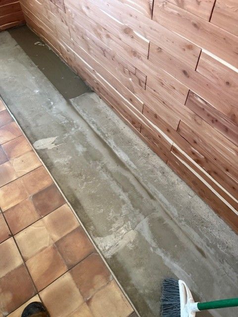 Wooden-paneled walls and tile floor meet a concrete strip. A broom is in the lower right.