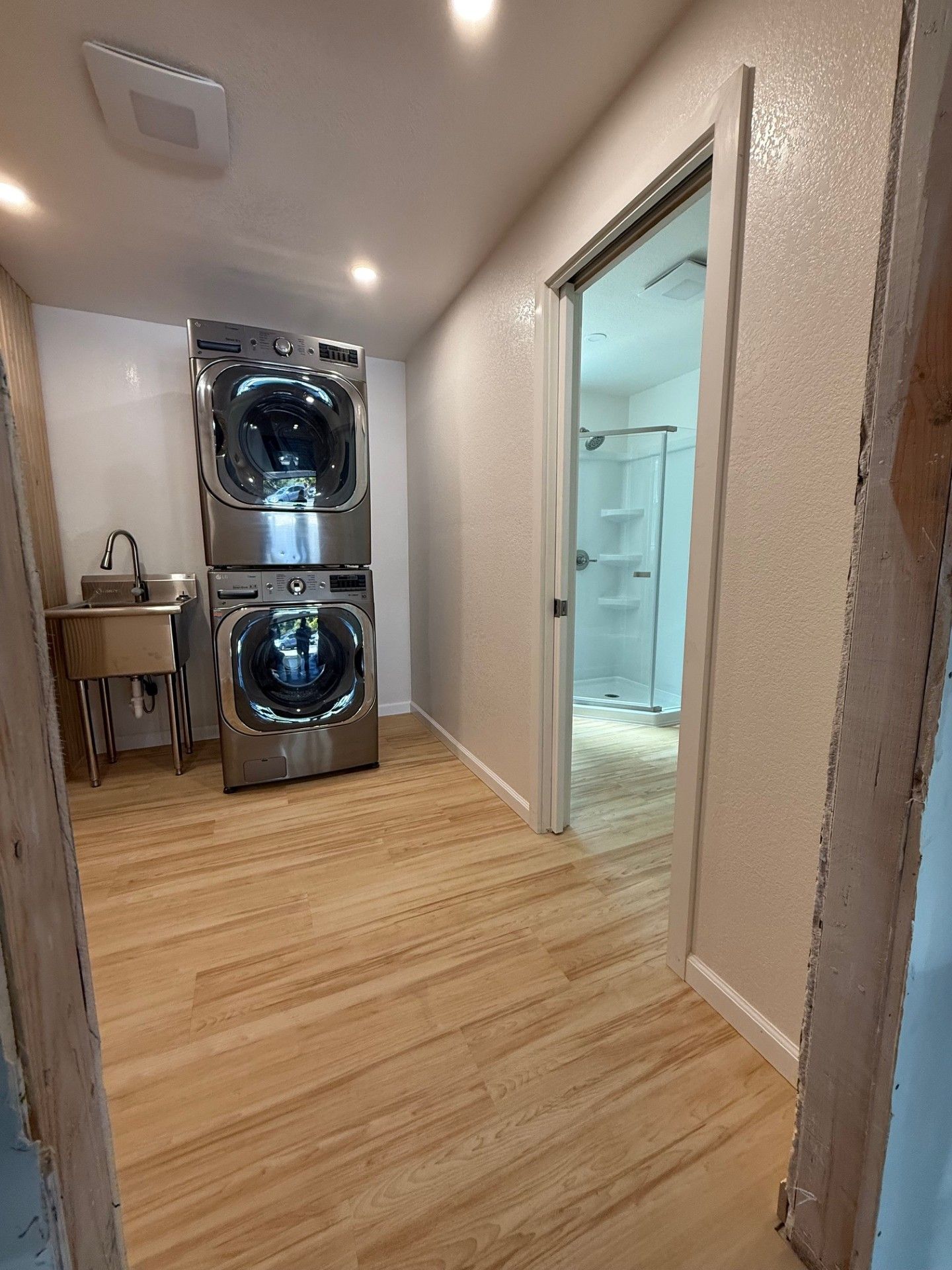 Laundry room with stacked washer/dryer, sink, and doorway to bathroom. Wooden floor, neutral walls.