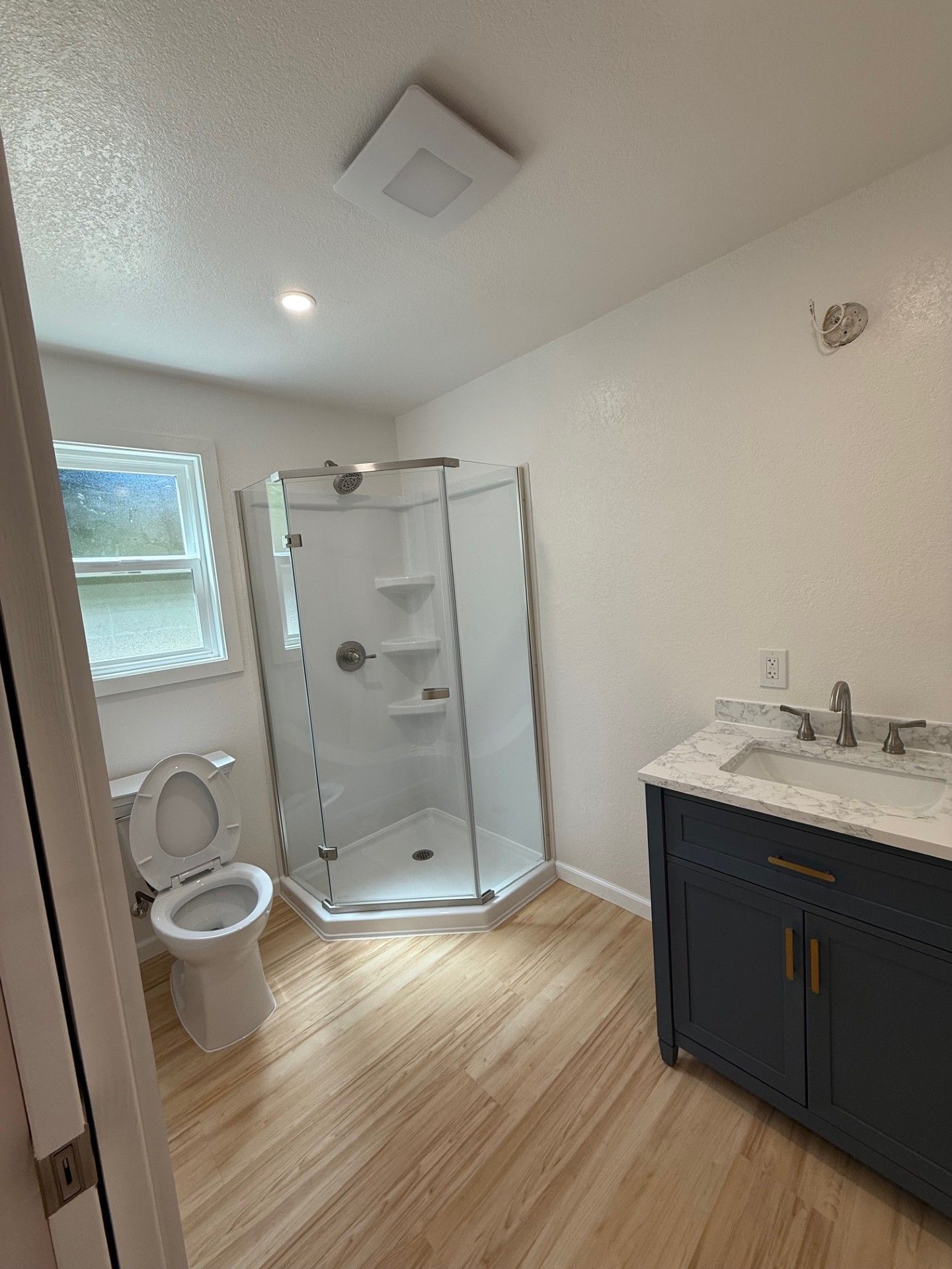 Bathroom with a shower, vanity, and toilet. Features white walls, light wood floor, and a dark blue vanity.