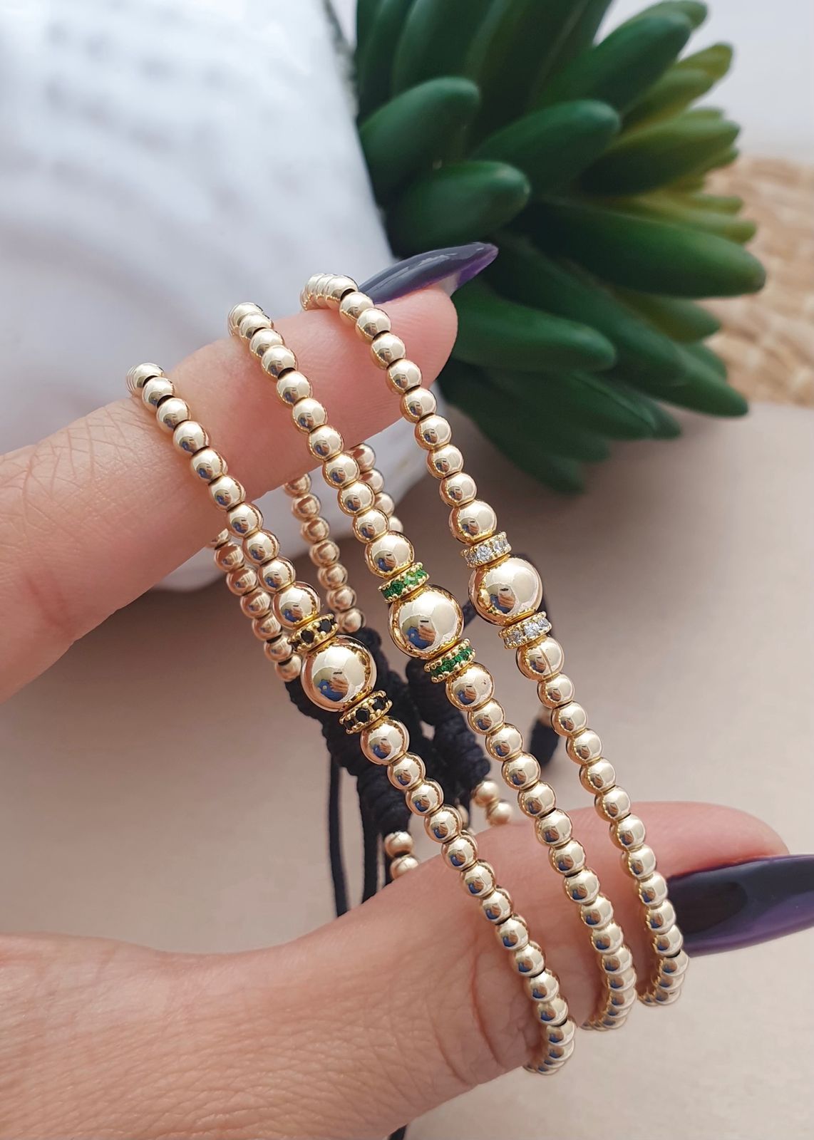 Tres pulseras ajustables con cuentas de oro, sostenidas por una mano, cada una con una cuenta central de oro