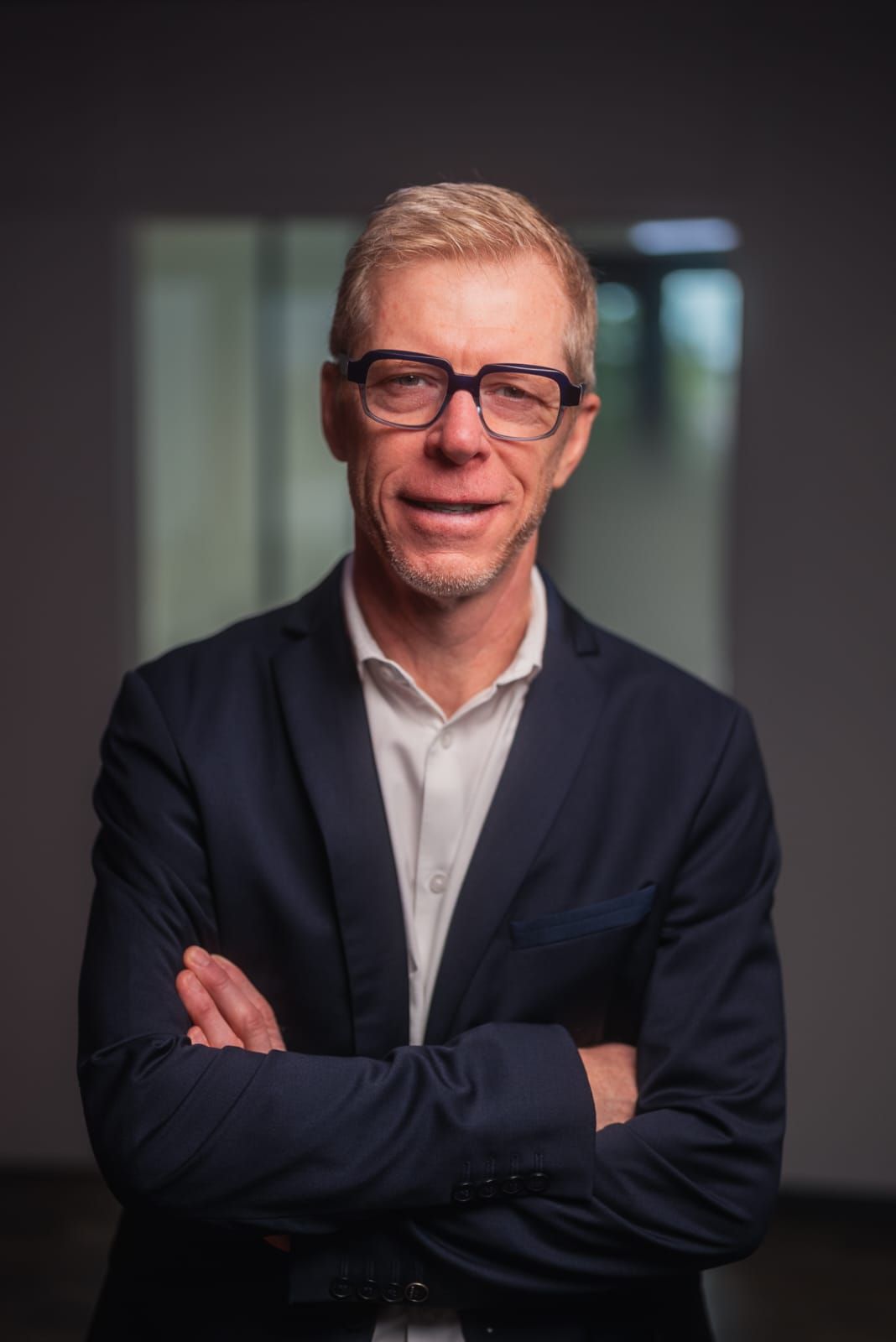 Un homme en blazer et lunettes, les bras croisés, sourit dans un espace moderne.