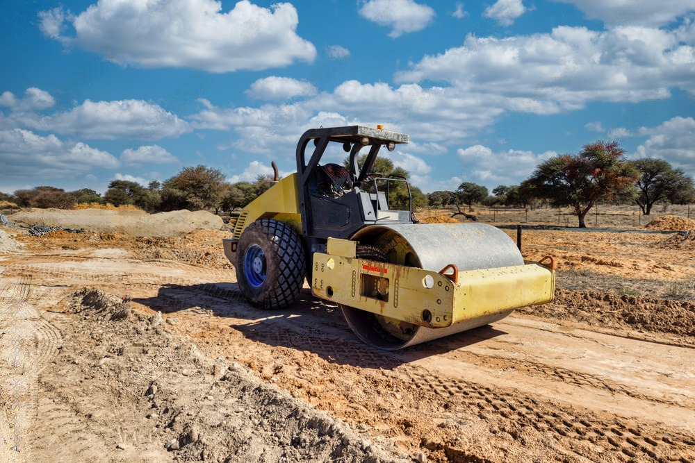 A yellow road roller is driving down a dirt road.