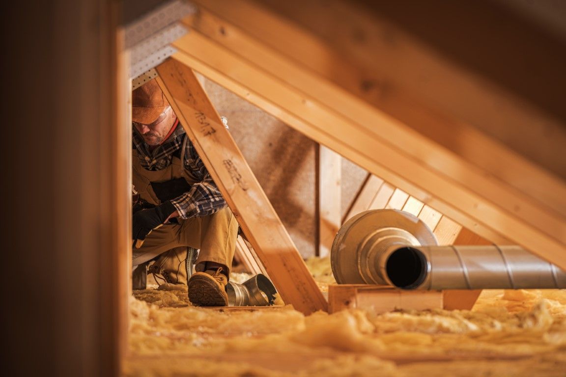 Worker inspecting attic insulation beside a silver duct in a wooden roof space