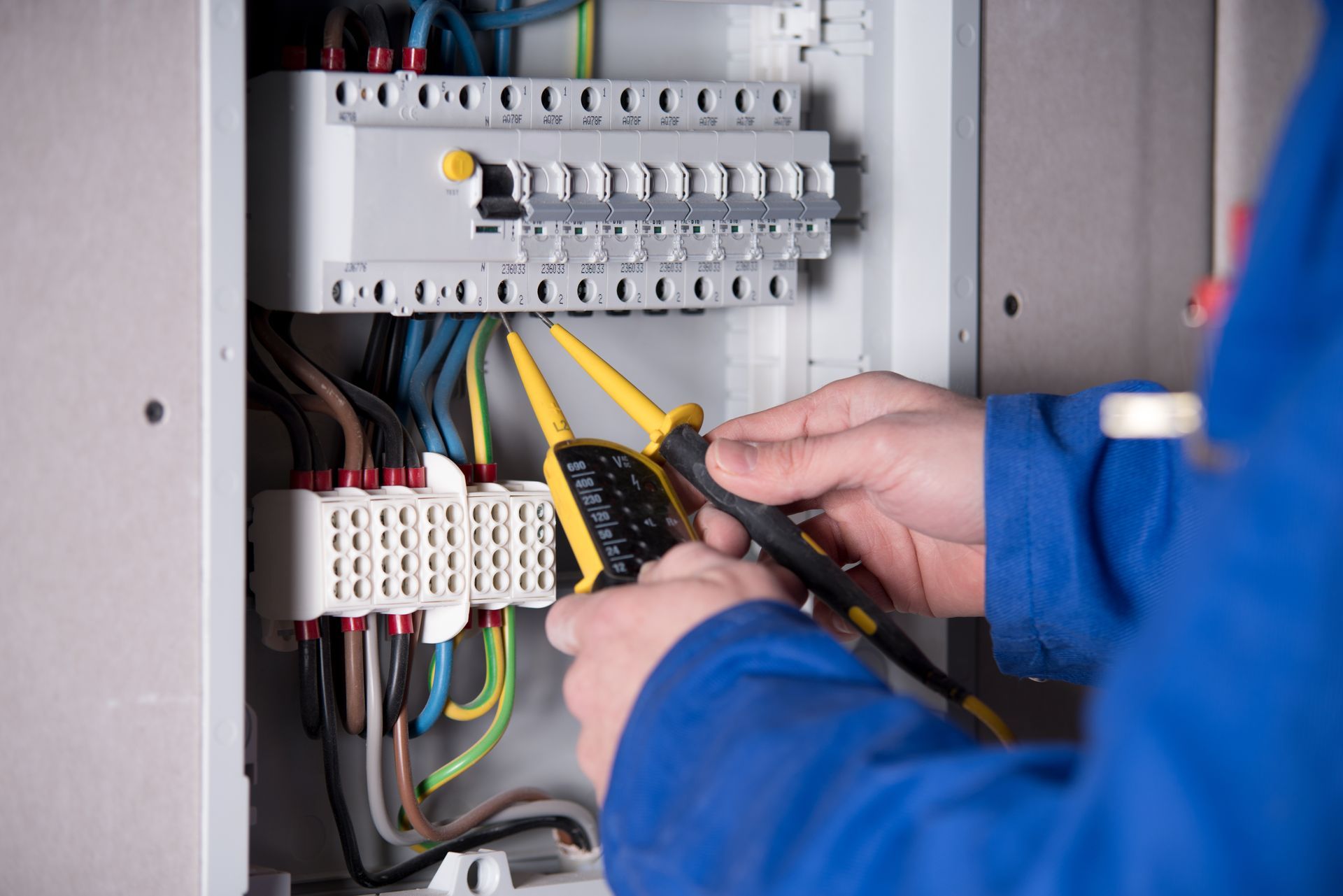 Elektriker in blauem Overall prüft einen Sicherungskasten mit einem Multimeter.