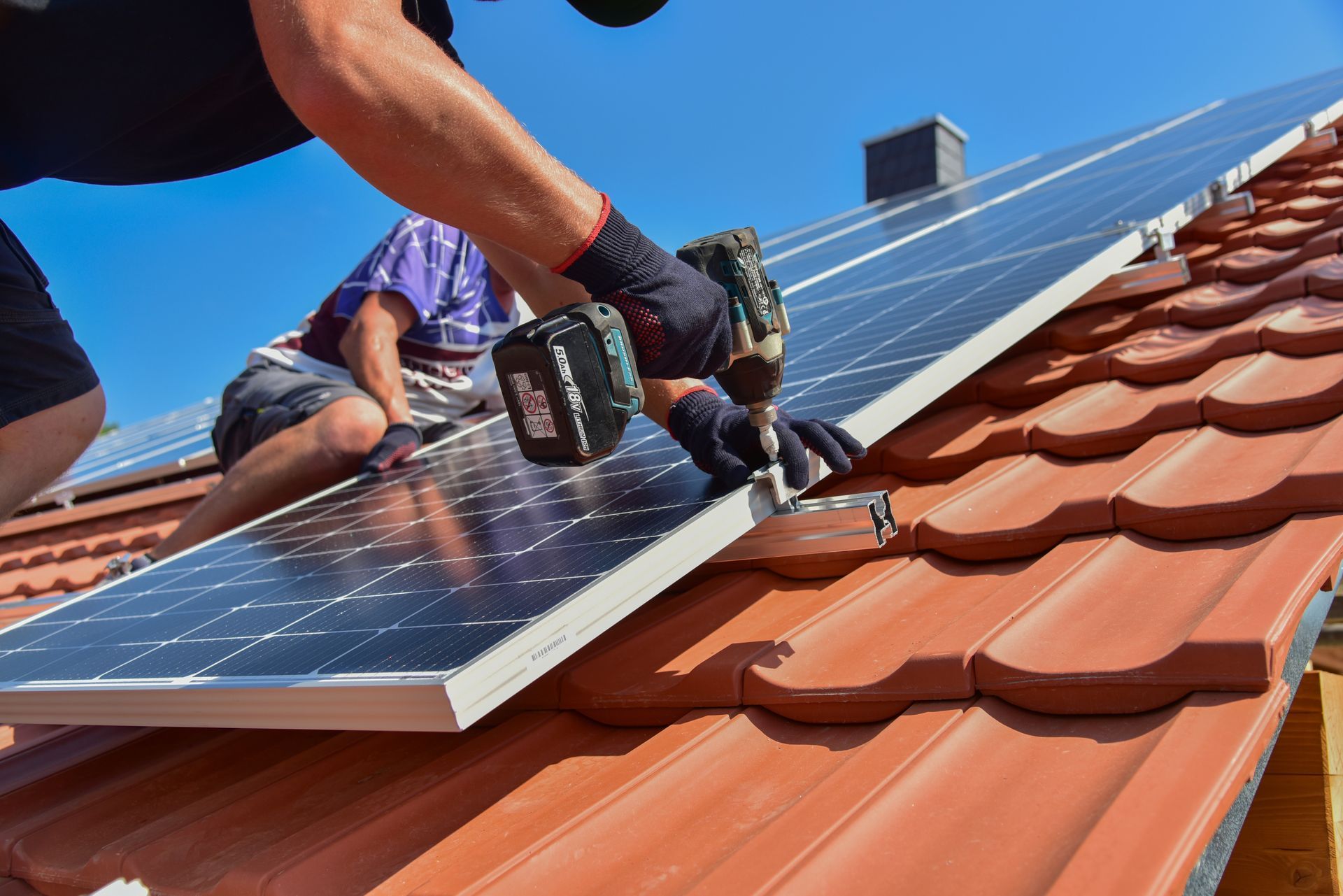 Zwei Personen montieren Solarpaneele auf einem roten Ziegeldach; eine davon benutzt eine Bohrmaschine.