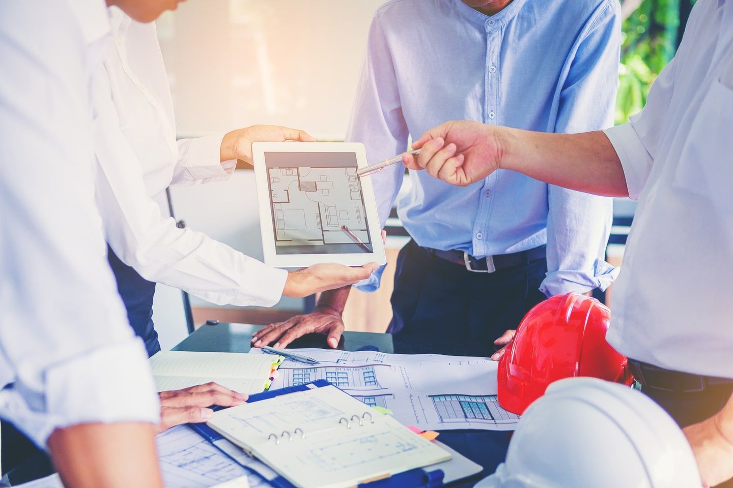 Menschen in einer Besprechung, die Baupläne auf einem Tablet prüfen, rote und weiße Schutzhelme auf einem Schreibtisch.