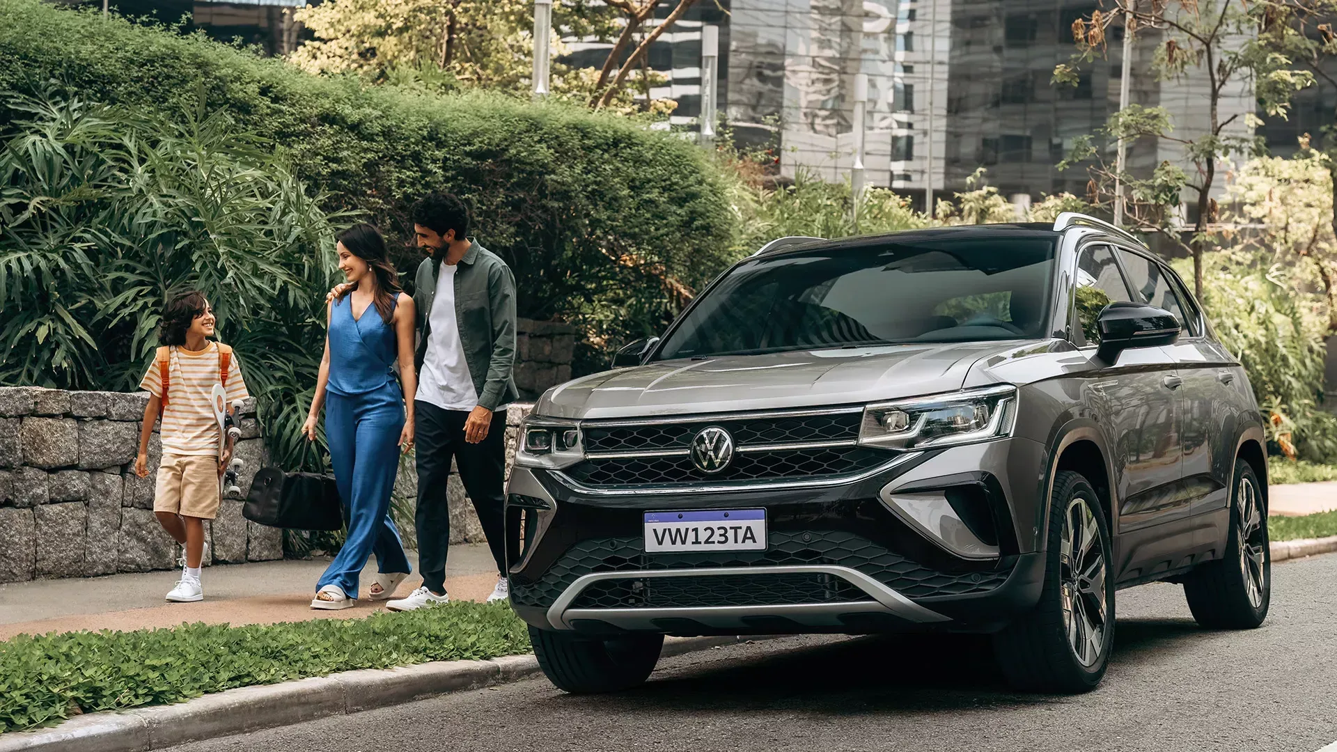 Uma família caminha perto de um SUV prateado estacionado na rua; o cenário urbano é visível ao fundo.