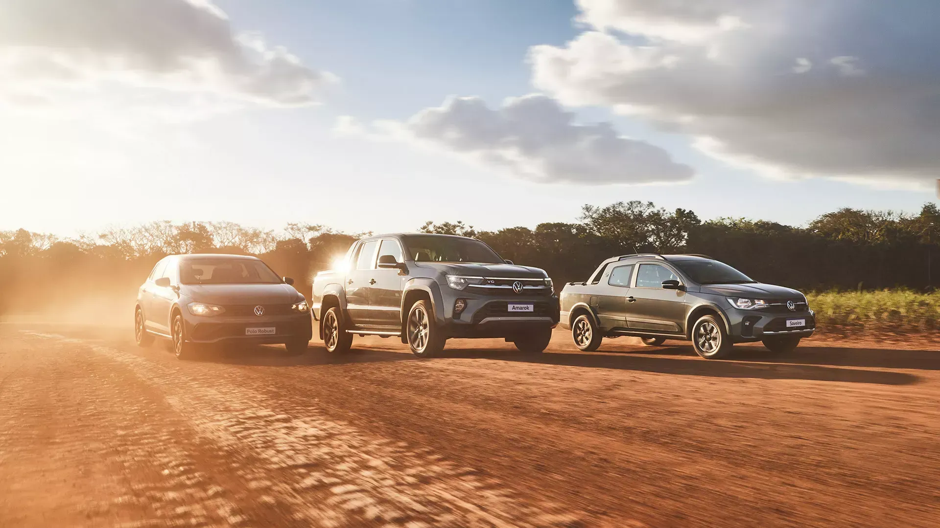 Três veículos trafegando por uma estrada de terra poeirenta sob um céu ensolarado.