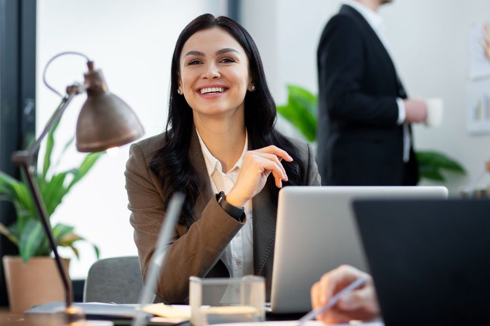 Smiling professional at a desk with a laptop in a bright office, wearing a brown blazer.