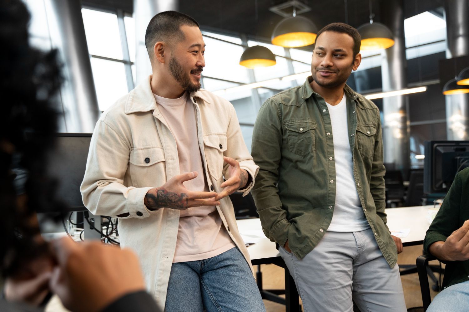 Two coworkers talking in a bright office, one gesturing while the other listens and smiles