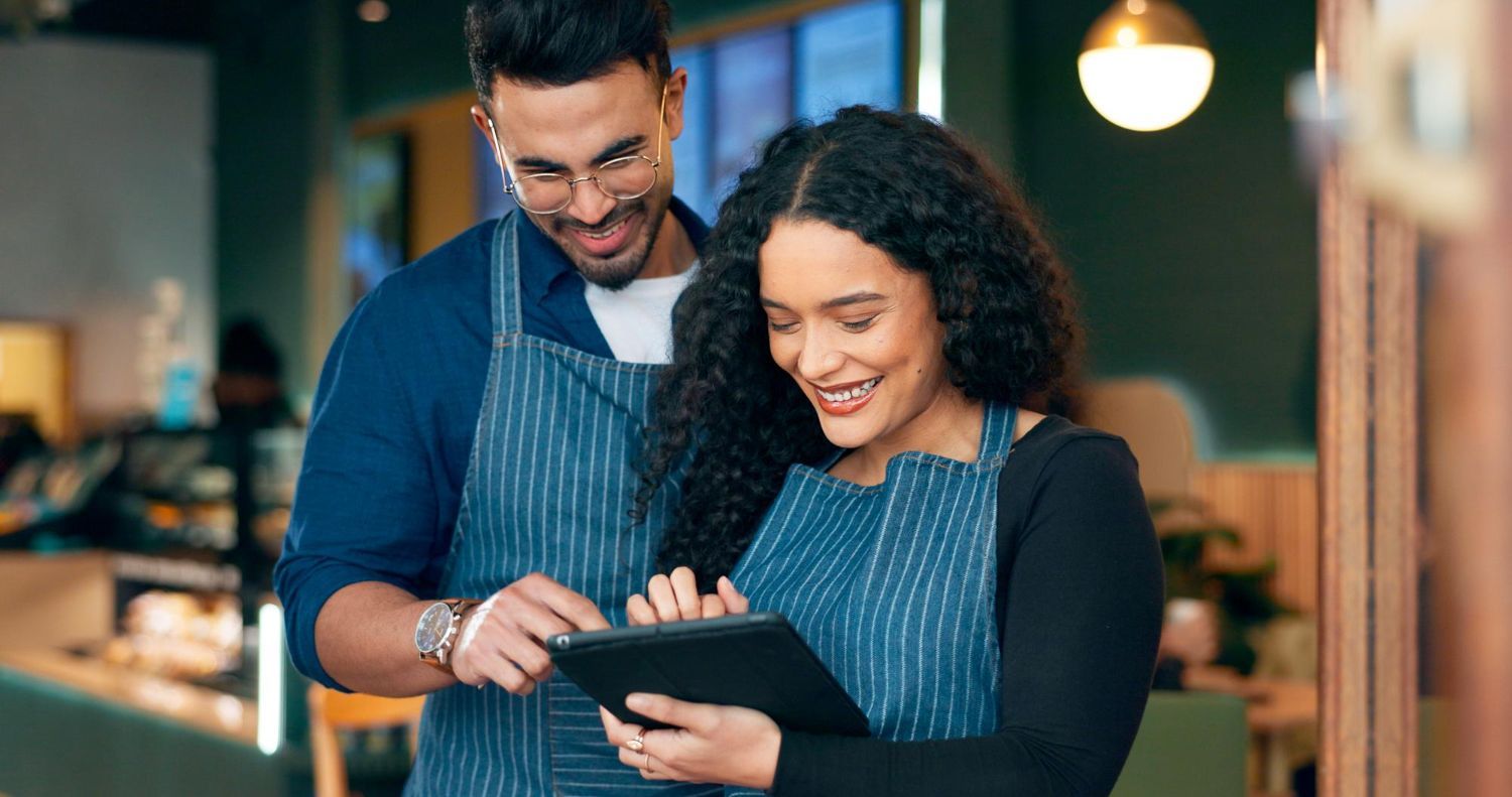 Two coworkers in aprons smiling and reviewing a tablet in a cafe.