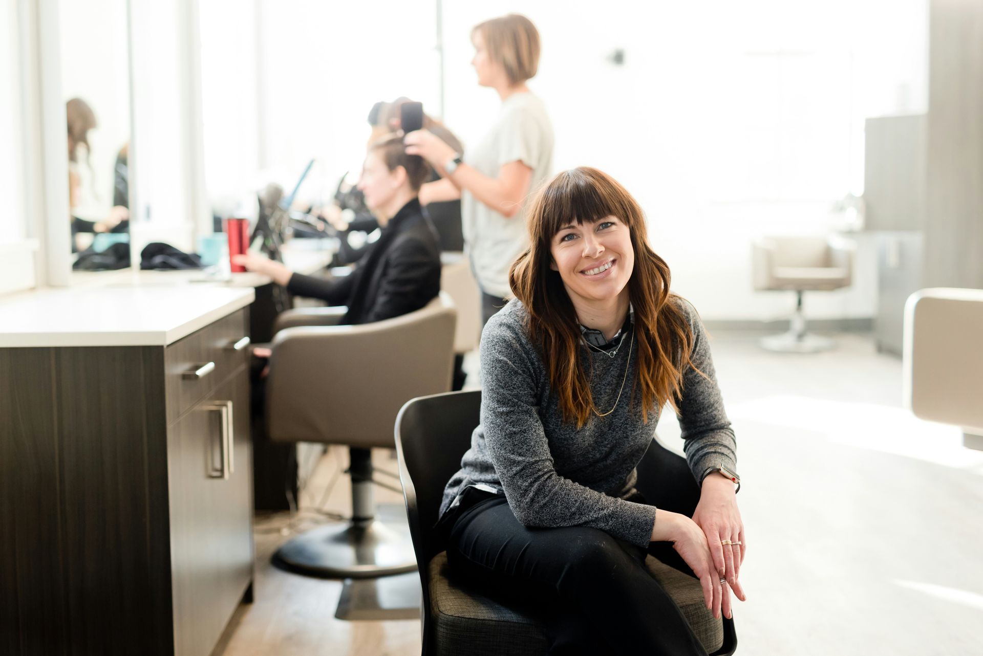 Woman smiling in a bright hair salon, with a stylist working on a seated client in the background