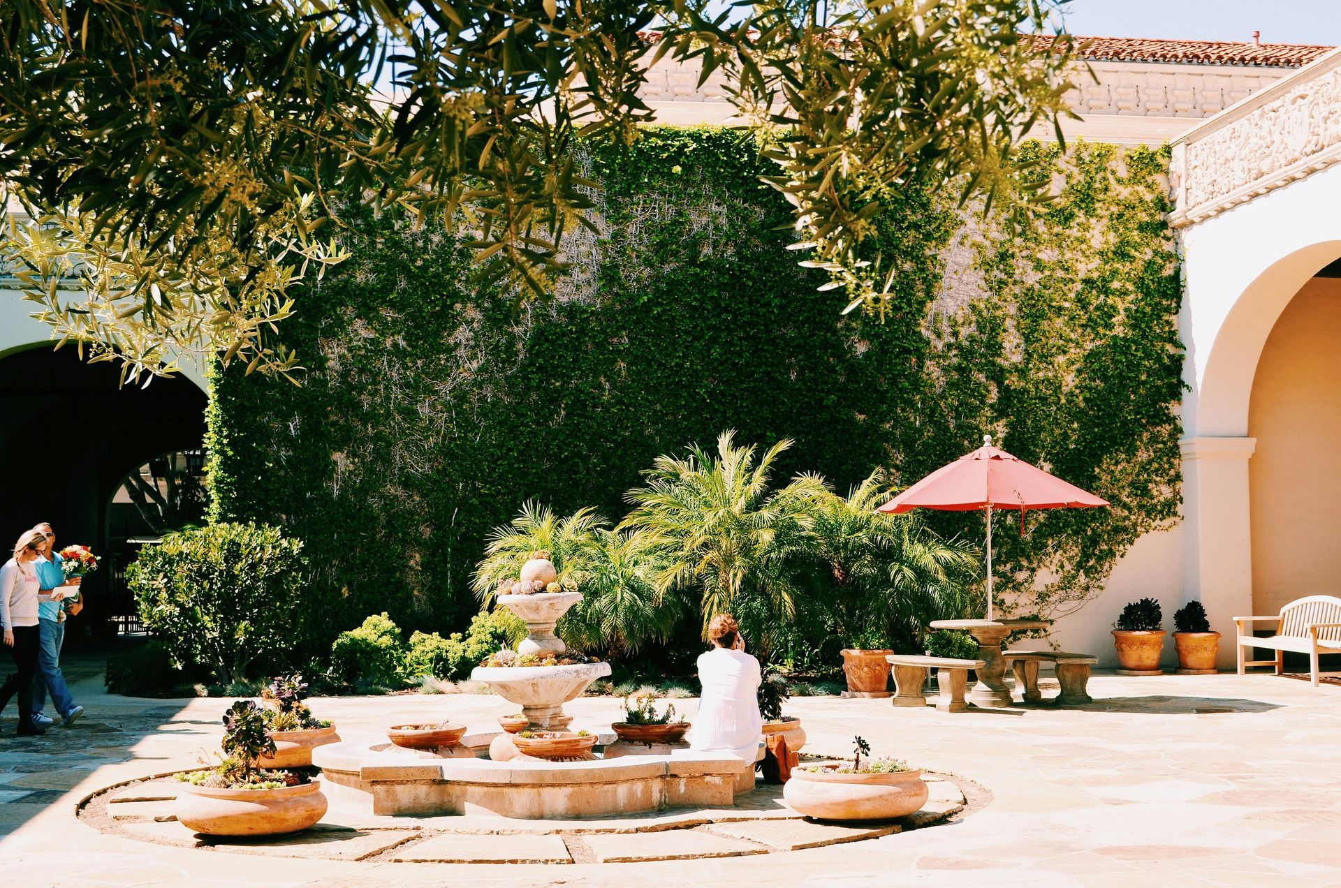 Sunny courtyard with tables, umbrellas, and lush greenery beside a white stucco building