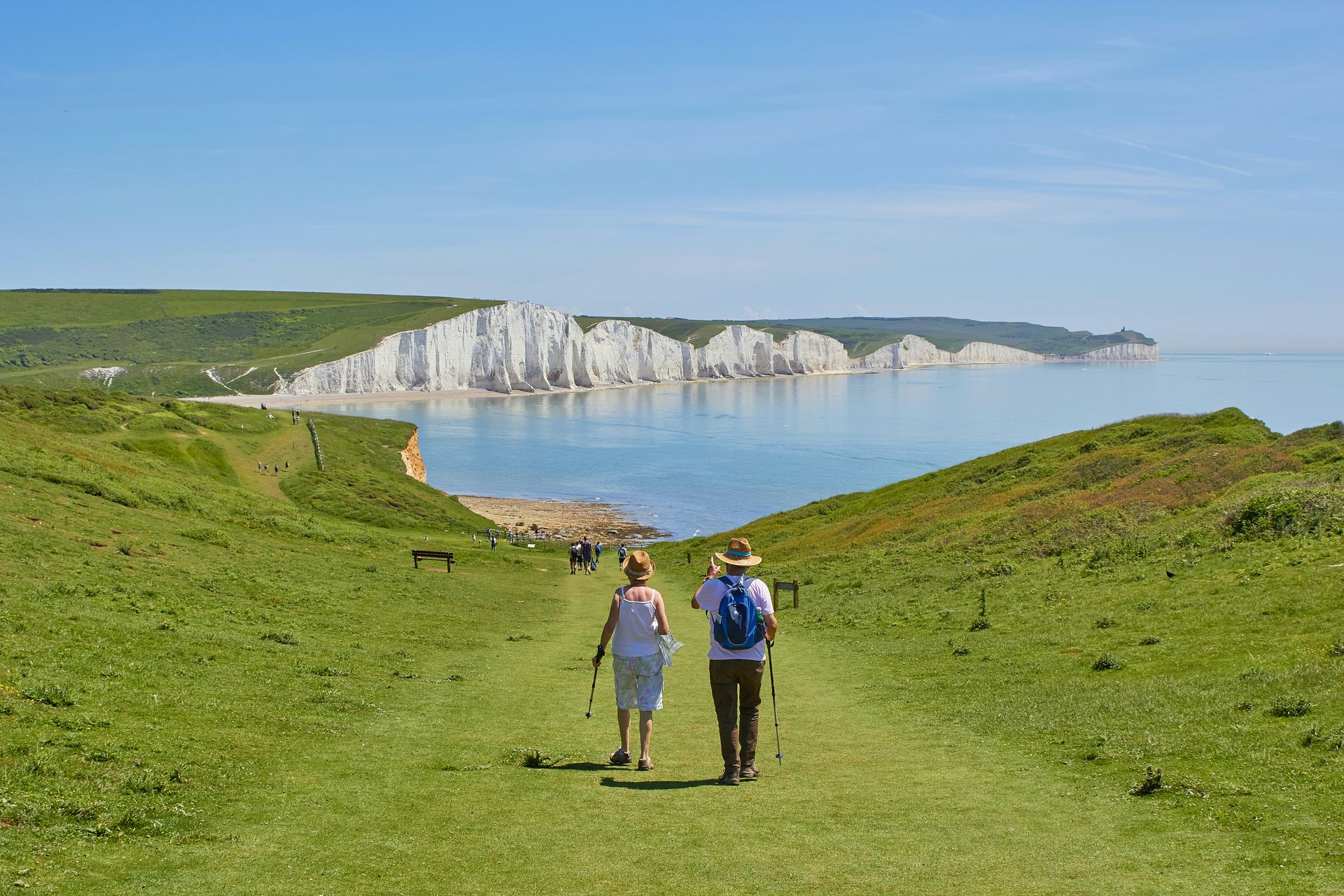 Two people walking on a grassy cliff path toward white sea cliffs and a calm blue bay
