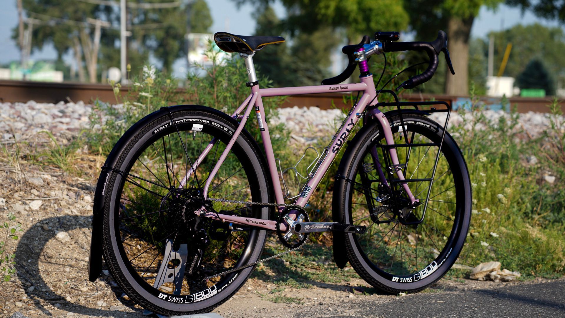 A pink bicycle is parked on the side of the road next to a train track.