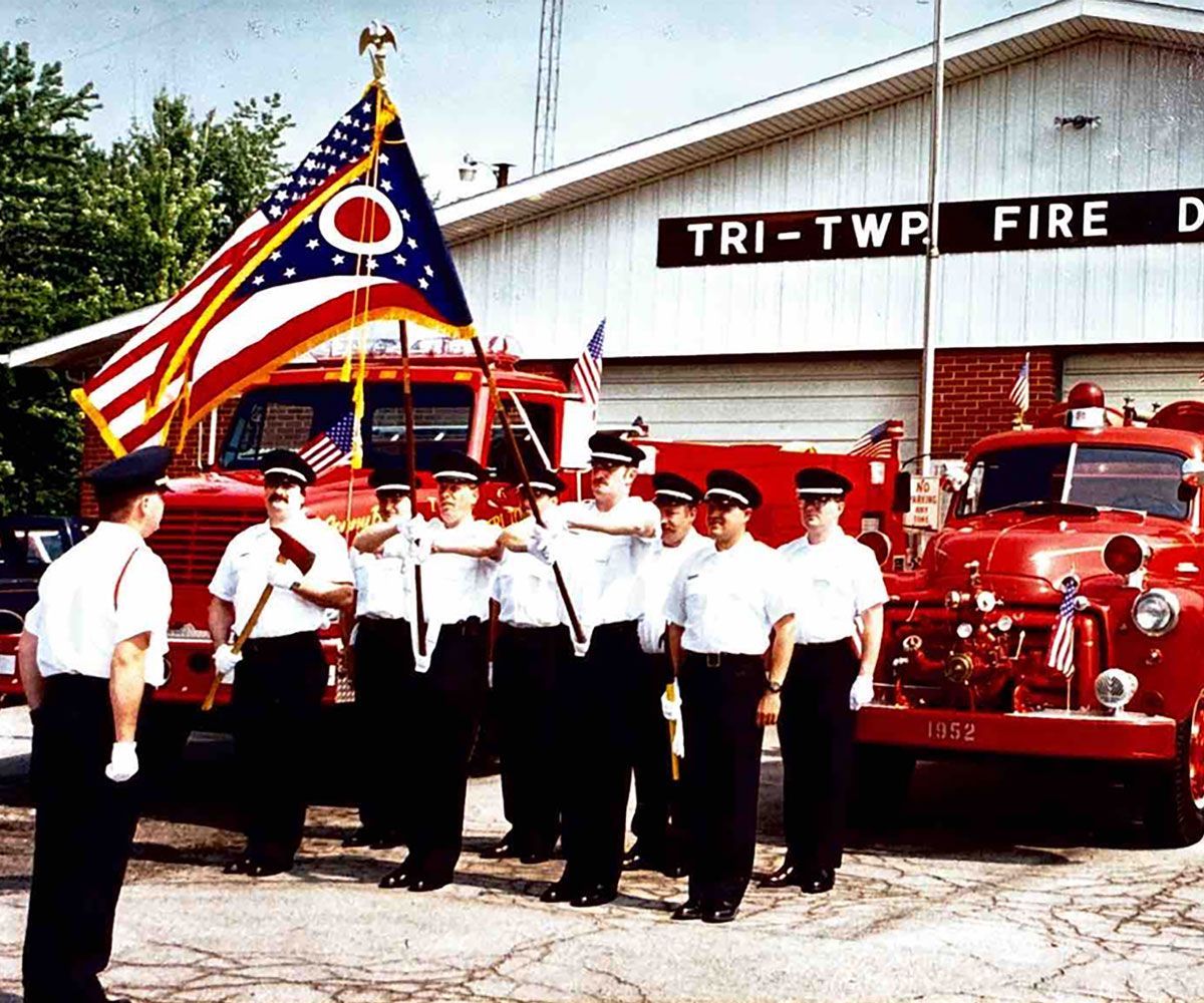 Tri Township historical photo of firemen standing at salute holding Ohio flag in front of original fire station circa 1970s or 1980s