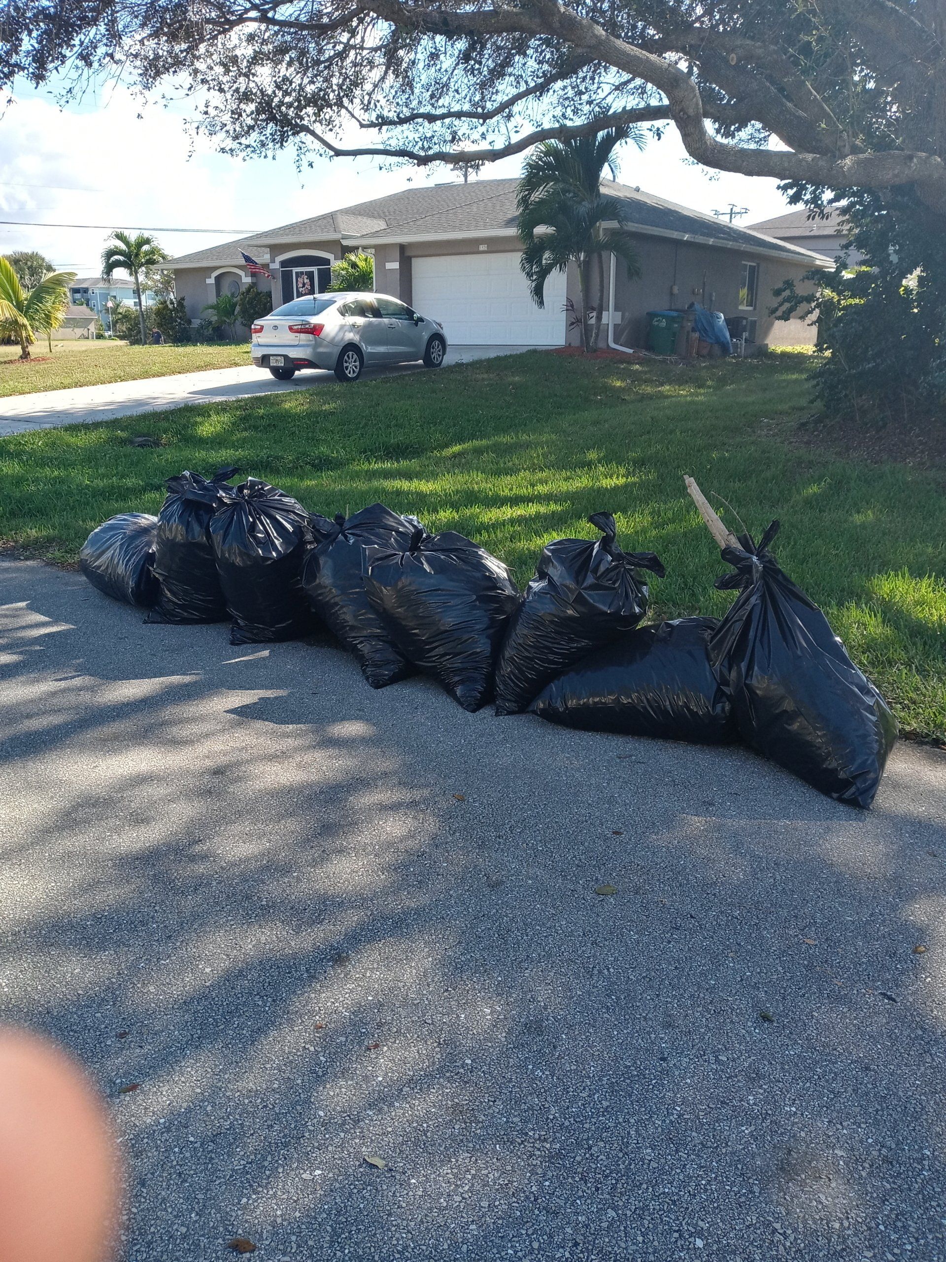 Black trash bags on a driveway in front of a suburban house with a car parked in the driveway.